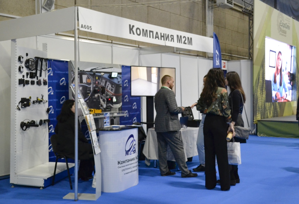 Стенд компании M2M на выставке, оформленный с демонстрацией оборудования для мониторинга и видеонаблюдения. Справа группа людей обсуждает продукцию с представителем компании, на заднем плане видно телевизор с рекламным видео и элементы выставочного зала. Стенд находится на синем ковре, на нем представлены компоненты для транспортных систем и оборудования для мониторинга.
